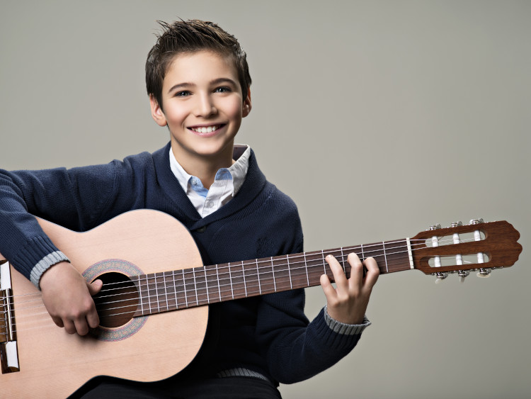 Featured image for Summer Recital 2019 - Music Lessons West depicting smiling teen boy in nice collared shirt and sweater playing a nylon string classical guitar.